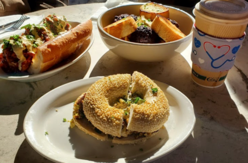 A plate with a sesame bagel sandwich, a bowl of golden bread, and a cup of coffee on a table.