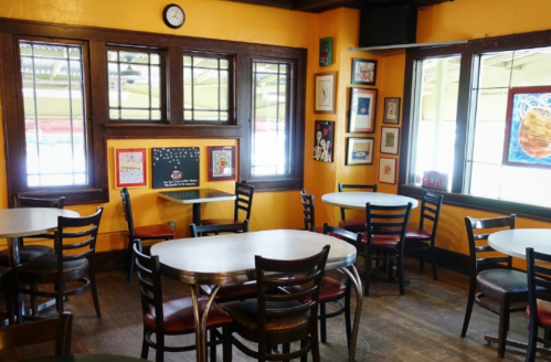 Cozy café interior with yellow walls, round tables, and black chairs, featuring artwork and large windows.
