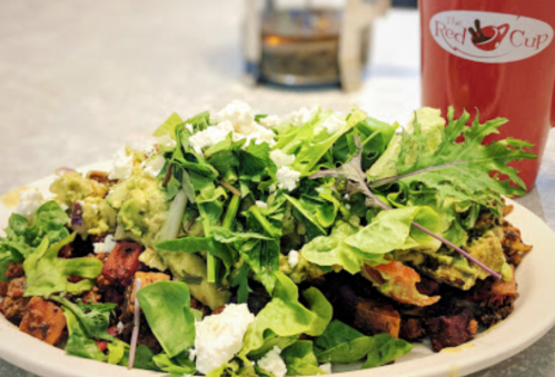 A colorful salad topped with greens, avocado, and feta cheese, served alongside a cup from The Red Cup café.