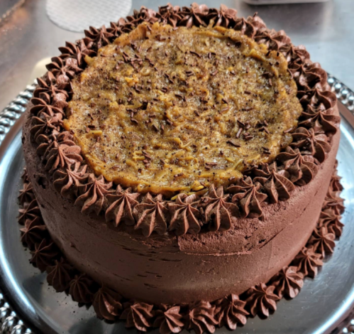 A round chocolate cake with a textured top, decorated with chocolate frosting and chocolate shavings around the edges.