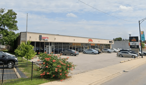 A single-story commercial building with multiple storefronts and a parking lot, surrounded by greenery and blue skies.