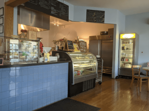 Cozy café interior with a display case, menu board, and refrigerators, featuring wooden tables and a warm atmosphere.