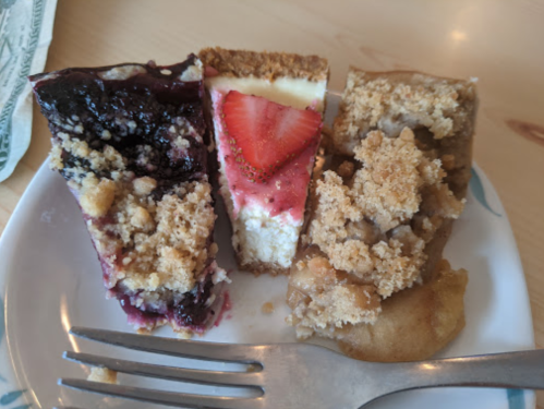 A plate with three slices of dessert: blueberry, strawberry cheesecake, and apple crumble, with a fork beside it.