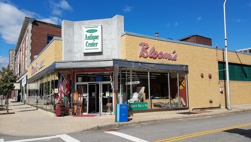 A vintage antique center with large windows, a sign reading "Bloomi's," and an American flag outside on a sunny day.
