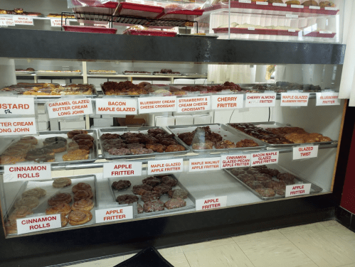 Display case filled with various pastries, including donuts, fritters, and croissants, labeled with their names.