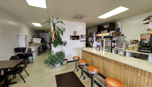 A small diner interior with a counter, orange stools, a plant, and a worker behind the counter. Boxes are stacked in the background.