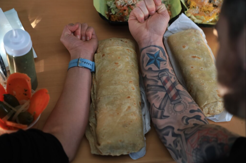 Two arms resting on a table, one with a watch and a tattoo, beside two large burritos.