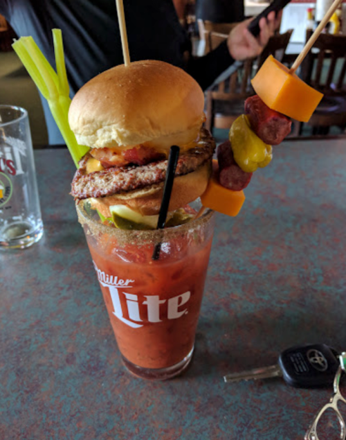 A tall glass of Bloody Mary topped with a mini burger and a skewer of sausage and cheese, garnished with celery.