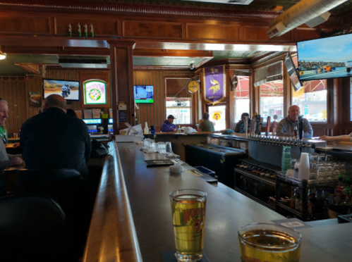 A bar scene with patrons seated, drinks on the counter, and multiple TVs showing sports in a cozy atmosphere.