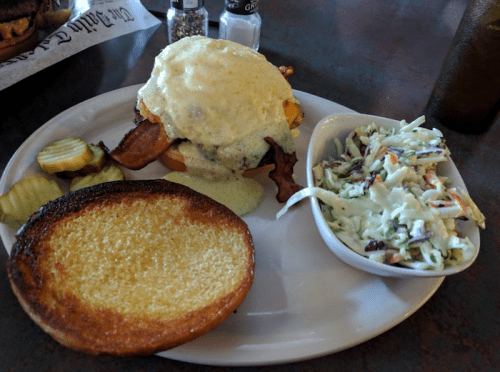 A plate featuring a sandwich with bacon and creamy sauce, alongside coleslaw, pickles, and a toasted bun.