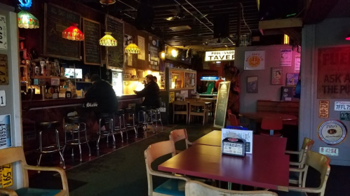 A cozy tavern interior with a bar, patrons seated, colorful lamps, and vintage decor. Tables are set for dining.