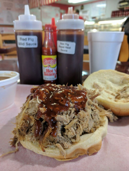 A pulled pork sandwich topped with barbecue sauce, accompanied by bottles of sauce and a cup of coleslaw.