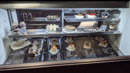 A display case filled with various desserts, including cakes, cupcakes, and pastries, each labeled with prices.