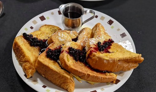 A plate of French toast topped with berry sauce and powdered sugar, accompanied by a small jug of syrup.