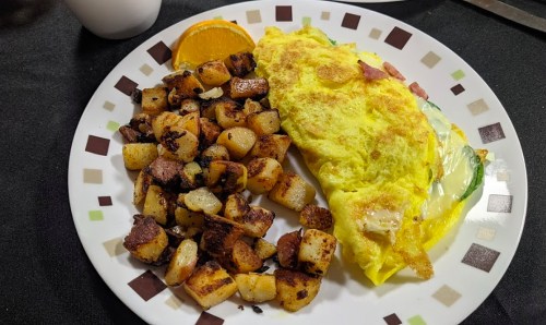 A plate with a fluffy omelette filled with vegetables and ham, alongside crispy golden-brown diced potatoes and an orange slice.