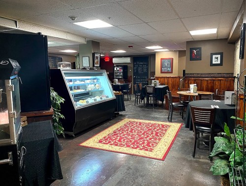 Cozy café interior with tables, a display case, and a decorative rug, featuring warm lighting and wooden accents.