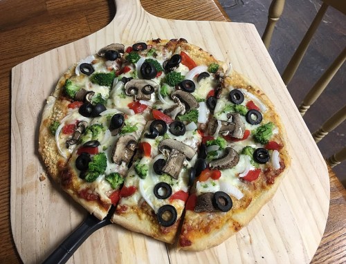 A freshly baked pizza topped with mushrooms, olives, broccoli, and red peppers, sliced on a wooden board.
