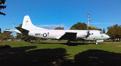 A retired Navy aircraft displayed on a grassy area, surrounded by trees and under a clear blue sky.