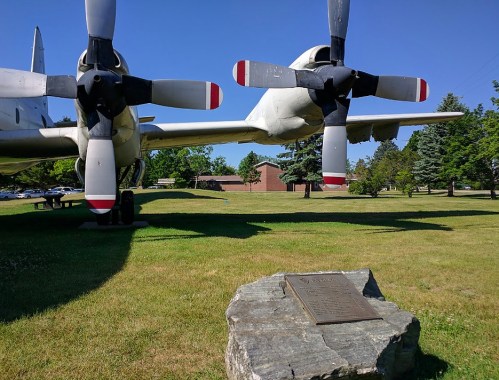 A vintage aircraft on display in a park, with a plaque on a rock in the foreground and trees in the background.