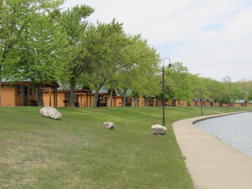 Row of wooden cabins along a grassy path by a calm lake, surrounded by trees and a cloudy sky.