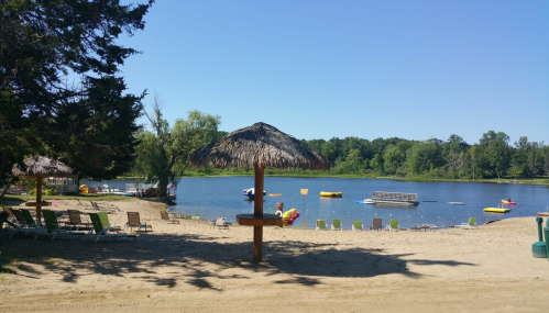 A sandy beach by a calm lake, featuring lounge chairs, a thatched umbrella, and colorful inflatable floats.