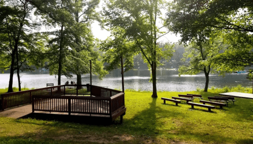 A serene lakeside scene with a wooden deck, green trees, and benches overlooking calm water.