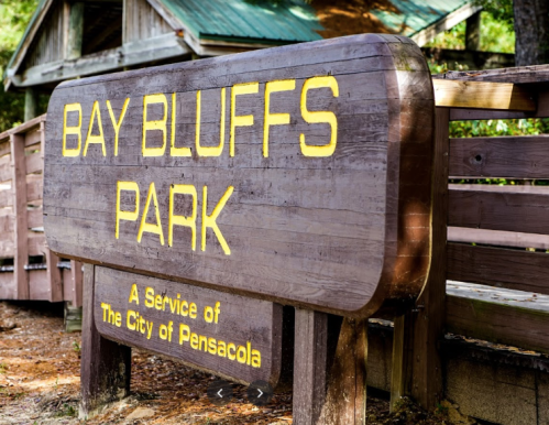 Sign for Bay Bluffs Park, featuring wooden design and yellow lettering, surrounded by trees and park structures.