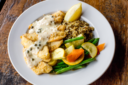 A plate of fried fish with caper sauce, served with rice and a mix of sautéed vegetables and lemon wedges.