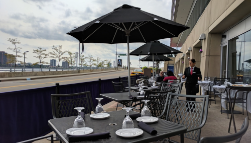 Outdoor dining area with tables set for meals, umbrellas, and a view of a street and waterfront. A waiter is present.