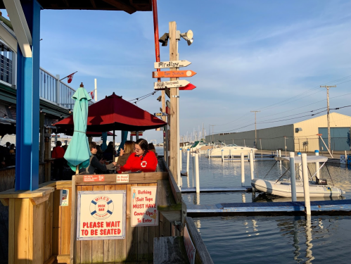 A waterfront restaurant with outdoor seating, umbrellas, and a sign directing to various locations, overlooking boats.