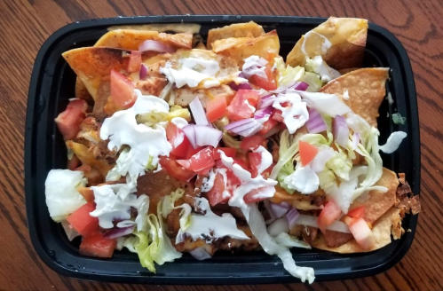 A black takeout container filled with nachos topped with cheese, tomatoes, lettuce, onions, and sour cream.
