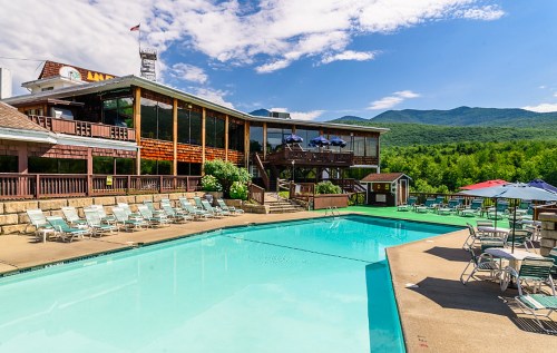 A scenic view of a pool area with lounge chairs, surrounded by mountains and a lodge with large windows.