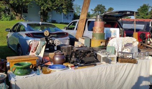 A table filled with vintage items, including a typewriter, jars, and various antiques, with cars parked in the background.