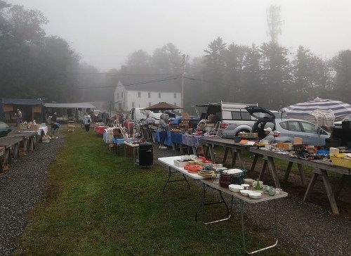 A foggy outdoor market with tables displaying various items, surrounded by trees and a few buildings in the background.