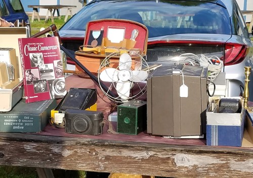 A table filled with vintage cameras, a fan, and various antiques, with a car in the background.