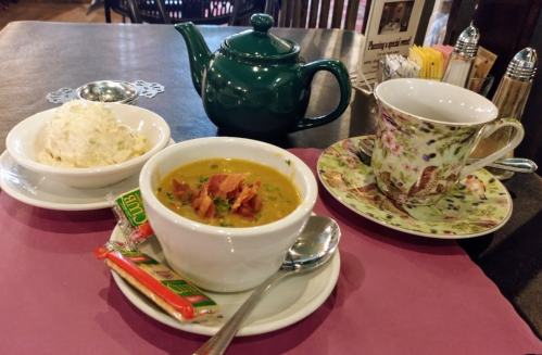 A green teapot, a floral teacup, a bowl of soup topped with bacon, and a side of creamy rice on a table.