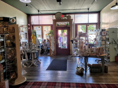 A cozy shop interior with displays of gifts and crafts, leading to a bright entrance with people seated outside.