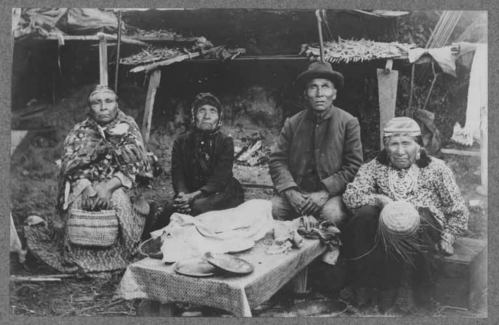Four individuals seated in front of a rustic shelter, surrounded by traditional items and a table with various objects.