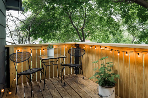 A cozy balcony with two chairs, a table, and string lights, surrounded by greenery and a potted plant.