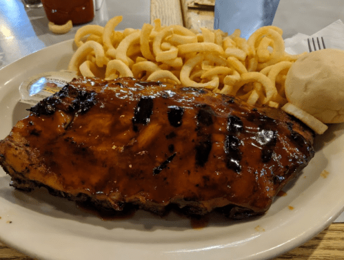 A plate featuring grilled ribs glazed with barbecue sauce, curly fries, and a soft roll.