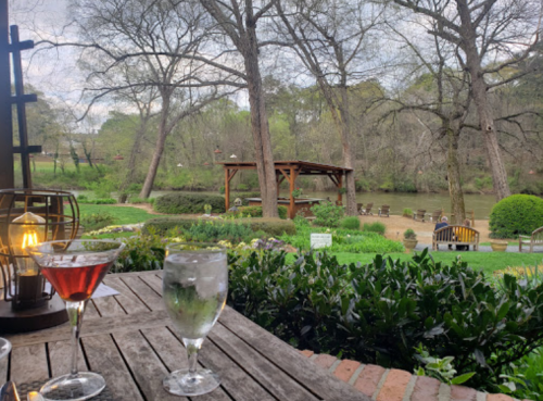 A serene outdoor scene featuring two cocktails on a wooden table, with a lush garden and river in the background.
