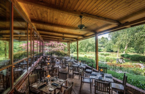 A cozy restaurant patio with wooden tables and chairs, surrounded by greenery and colorful flowers.