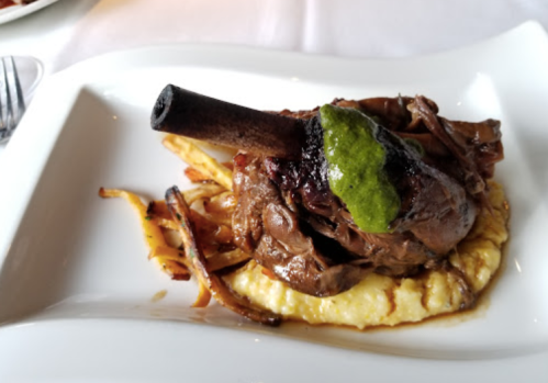 A plated dish featuring a lamb shank on creamy polenta, topped with green sauce and accompanied by crispy fries.
