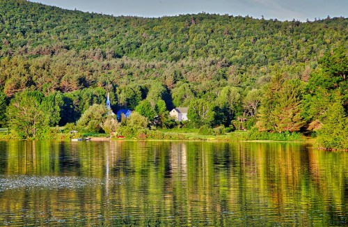 A serene lake reflects lush green trees and a quaint house nestled among the hills.