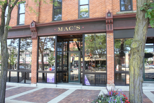Exterior view of a brick building with large glass windows, featuring a sign that reads "MAC'S." Trees and flowers in foreground.