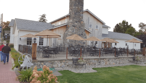 A rustic restaurant with a stone chimney, outdoor seating, and umbrellas, surrounded by greenery and a brick pathway.