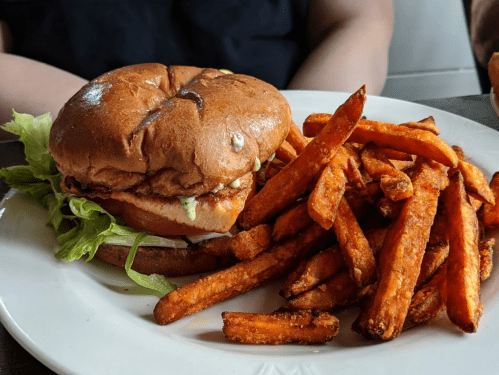 A brown bun sandwich with lettuce and a chicken patty, served with a side of crispy sweet potato fries.
