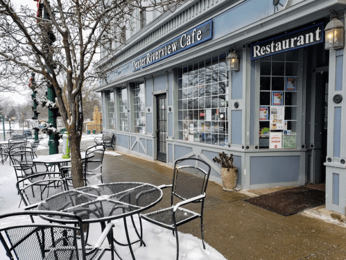A snowy scene outside the Dexter Riverview Cafe, featuring tables and chairs on the sidewalk near the entrance.