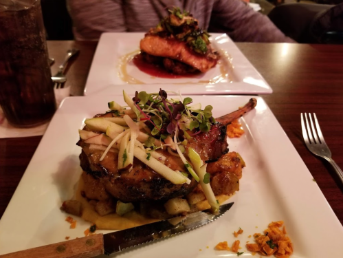 A plated pork chop topped with apple slices and microgreens, served with a side of diced fruit and vegetables.