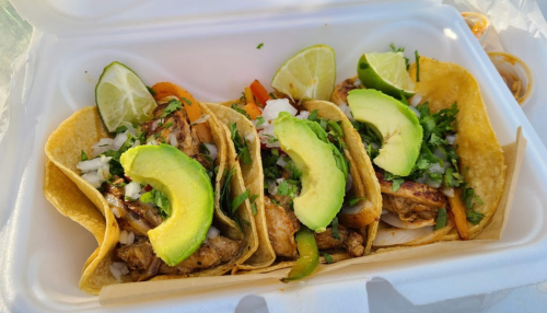 Three tacos filled with grilled chicken, onions, cilantro, avocado slices, and lime wedges in a takeout container.